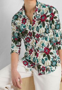 Hvid skjorte med knaplukning, der har et levende blomsterprint i rød, grøn og blå, med oprullede ærmer og krave. Letvægtsstof.