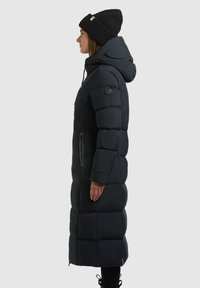 Dolga črna puffer jakna s kapuco, s prešitim dizajnom, z zip žepom na strani in logotipom na ovratniku. Nosena z črnim beaniejem.