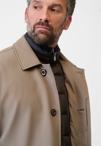 Beige yttre kappa med en slät textur, med ett svart innerfoder i puffer-material, en krage och två knappar. Innehåller en sidoficka.