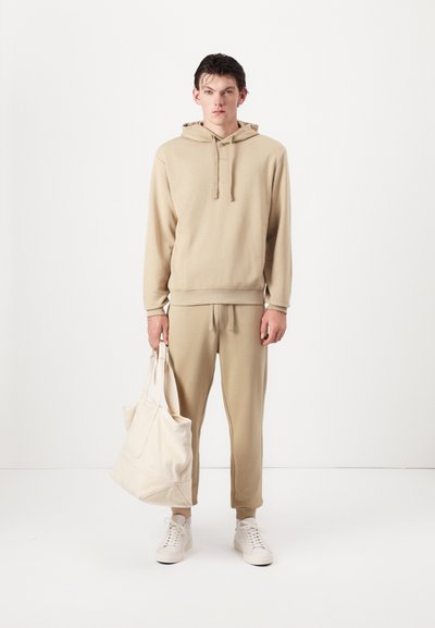Hoodie beige avec cordons de serrage, joggers beige assortis et baskets blanches ; tenant un sac fourre-tout de couleur claire. Texture de tissu lisse.