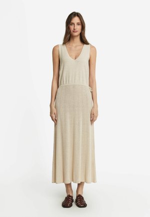 Beige stickad maxikjol med V-ringning, ärmlös design, smal midja och en flödande kjol. Matchad med mörkbruna sandaler.