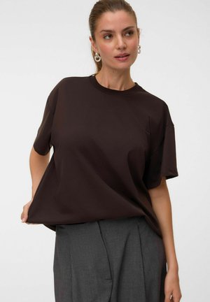 Femme aux cheveux châtain clair attachés en queue de cheval, porte une chemise ample marron foncé à manches courtes et un pantalon gris, regardant légèrement vers la droite.