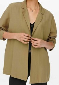 Persona che abbottona un blazer leggero di colore marrone chiaro sopra una maglietta nera con grandi bottoni e indossa una delicata collana con un pendente scuro.