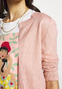 Ljust rosa kofta med ett strukturerat mönster som är lager över en vit t-shirt med grafisk design. Guldhalsband om halsen.