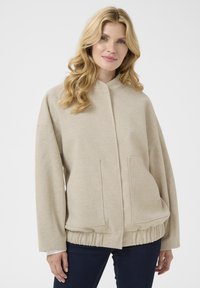 Sweat-shirt beige en tissu texturé, avec un col montant, deux poches avant et un ourlet resserré. Coupe décontractée avec des épaules tombantes.