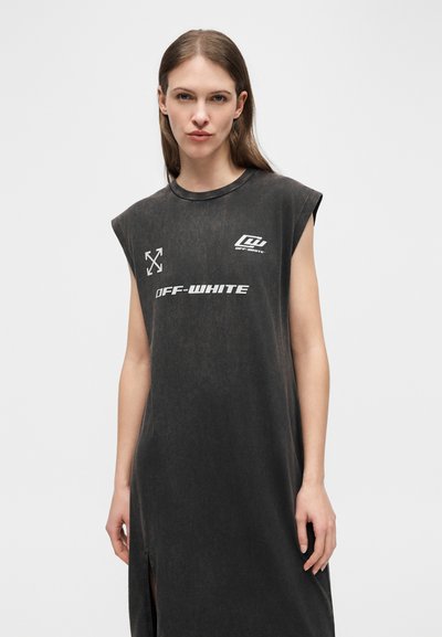 Γυναίκα με μακριά μαλλιά που φοράει μαύρο φόρεμα χωρίς μανίκια με λευκά λογότυπα "Off-White" στο μπροστινό μέρος, στέκεται μπροστά σε ένα απλό ανοιχτό φόντο.
