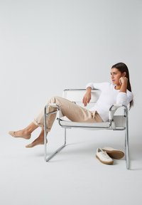Fauteuil lounge en chrome avec un siège en cuir blanc. Personne assise en pantalon crème et haut blanc, avec des ballerines beiges, des baskets beiges au sol.