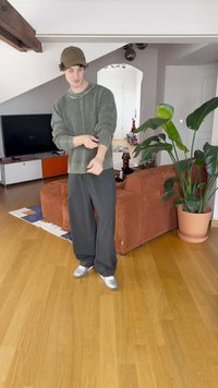 Green textured sweater, relaxed black trousers, and silver shoes. Modern interior with a brown corduroy couch and large indoor plant.