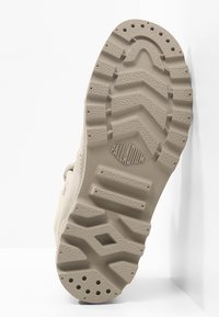 Semelle de chaussure en caoutchouc beige présentant un motif de bande de roulement en zigzag et un logo en relief. Surface texturée avec des trous de drainage pour la traction et la durabilité.