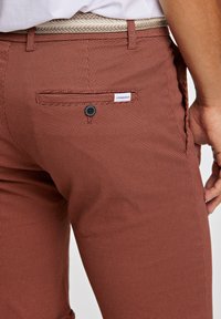 Pantalones de rayas rojas con una tela texturizada, que presentan un único bolsillo trasero con cierre de botón y una etiqueta situada encima.