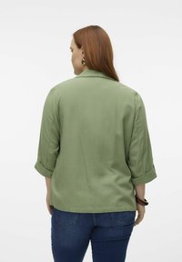 Chemise col vert en tissu lisse, avec des manches retroussées et une coupe décontractée. Associée à un jean bleu foncé.