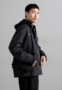 Casaco cinza escuro com fecho éclair e capuz, apresentando um corte descontraído, punhos e bolsos laterais com fecho. Tecido texturizado com um design minimalista.