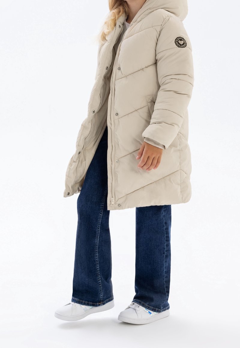 Enfant portant une doudoune beige à capuche, un jean bleu et des baskets blanches, debout sur un fond blanc.