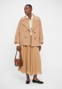 Beige wool double-breasted coat with large lapels, paired with a light top, flowing tan pleated skirt, black flats, and a brown shoulder bag.