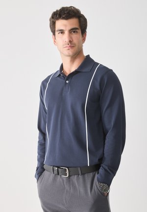 Polo a maniche lunghe di colore blu nautico con strisce bianche sui lati, colletto e chiusura con bottoni. Tessuto morbido in misto cotone. Indossato con pantaloni grigi.