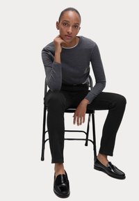 Γκρι μπλούζα με μακριά μανίκια, μαύρο skinny παντελόνι, γυαλιστερές μαύρες loafers. Μοντέλο καθισμένο σε μαύρη αναδιπλούμενη καρέκλα με φόντο λευκό.