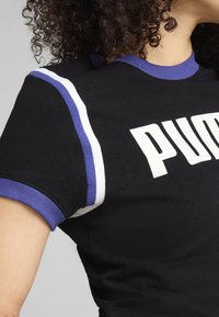 Camiseta negra con detalles a rayas blancas y moradas en las mangas. Presenta un gran logo blanco de "PUMA" en la parte frontal. Material de algodón.