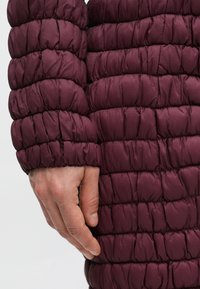 Bordeauxrote Steppjacke mit einem gesteppte Muster; Nahaufnahme einer Hand, die auf dem Ärmel ruht; glatter, glänzender Stoff mit elastischen Bündchen.