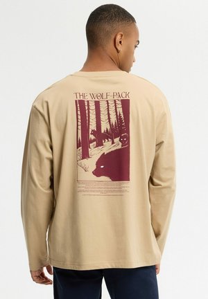 Hombre que lleva una camisa de manga larga beige con un gráfico de lobos en un bosque en la espalda y el texto "The Wolf-Pack" sobre la imagen.