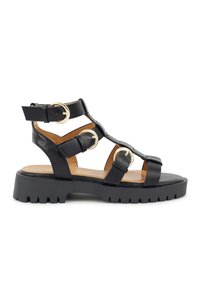 Schwarze Ledersandalen mit dicken Riemen, goldfarbenen Hardware-Akzenten und einer robusten Profilsohle. Offenes Design mit verstellbarem Knöchelriemen.