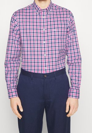 Homme portant une chemise à manches longues à carreaux rose et bleu boutonnée, rentrée dans un pantalon bleu foncé sur un fond blanc.