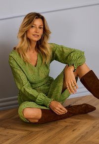 Frau mit blondem Haar sitzt auf Holzboden, trägt strukturiertes grünes Kleid und braune kniehohe Stiefel, mit goldenen Ringen und Ohrringen.