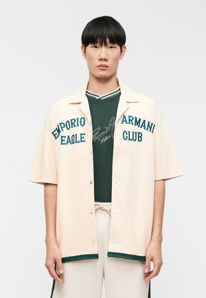 Άνδρας που φοράει μπεζ κοντομάνικο πουκάμισο με κουμπιά και κείμενο "Emporio Armani Eagle Club" πάνω σε σκούρο πράσινο τοπ και μπεζ παντελόνι με κορδόνια.