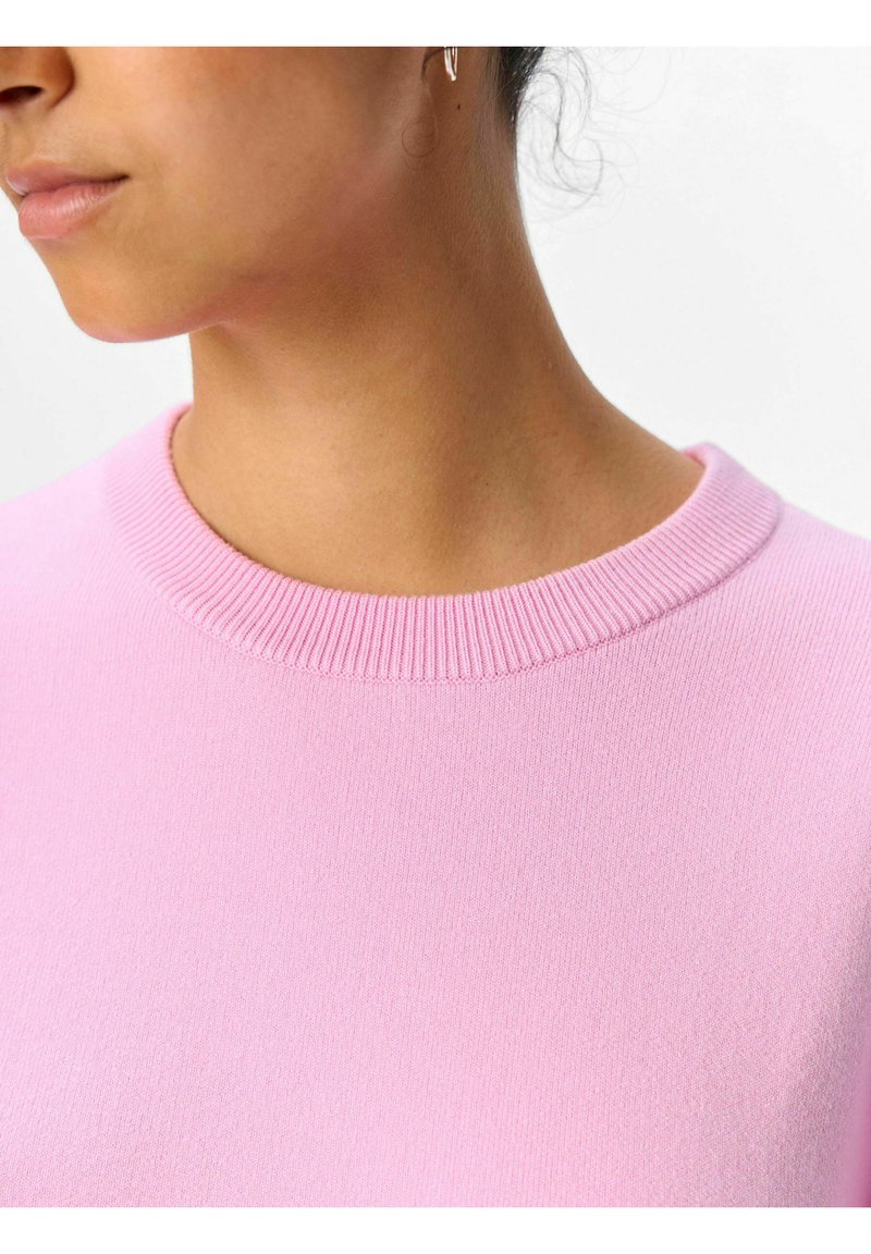 Jasnoróżowy sweter z prążkowanym okrągłym dekoltem, prezentujący miękką teksturę oraz zbliżenie na dekolt i obszar ramion.