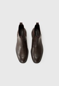 Botas de tobillo de cuero marrón con paneles laterales elásticos, punta redondeada y tiradores; textura suave con un ligero brillo, diseño minimalista.