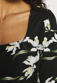 Czarna tkanina z białym kwiatowym nadrukiem przedstawiającym lilie. Wzór zawiera zielone łodygi oraz delikatny połysk, z kwadratowym dekoltem.