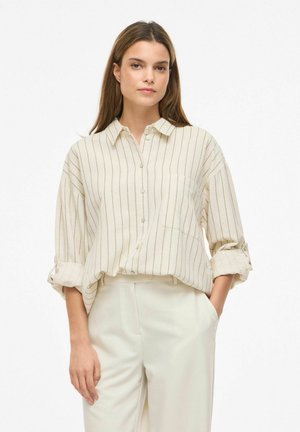 Jeune femme debout, portant une chemise écrue boutonnée avec de fines rayures verticales et un pantalon clair assorti.
