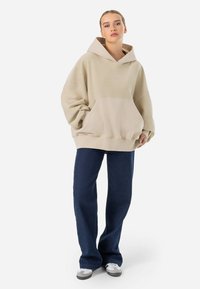 Felpa beige oversize con tasca frontale e cappuccio con cordini, abbinata a jeans a gamba larga blu scuro e sneaker chiare.