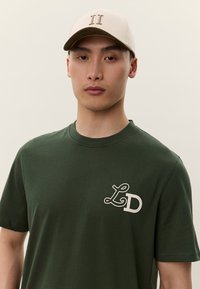 Tmavě zelené bavlněné tričko s bílým logem "LD" na levé straně hrudi, doplněné béžovou čepicí a tmavším kšiltem.