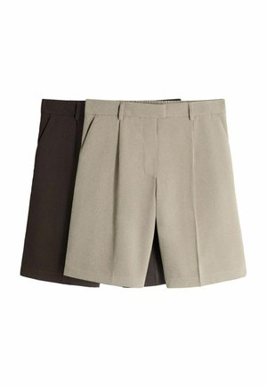 Kobieta w białym topie na ramiączkach, czarnym pasku, brązowych szortach do kolan i czarnych sandałach na obcasie stojąca na tle jednolitego białego tła.