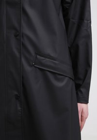 Svart vattentät regnrock med glansig yta. Har knapplås fram och en sidoficka. Slät textur och knälång design.