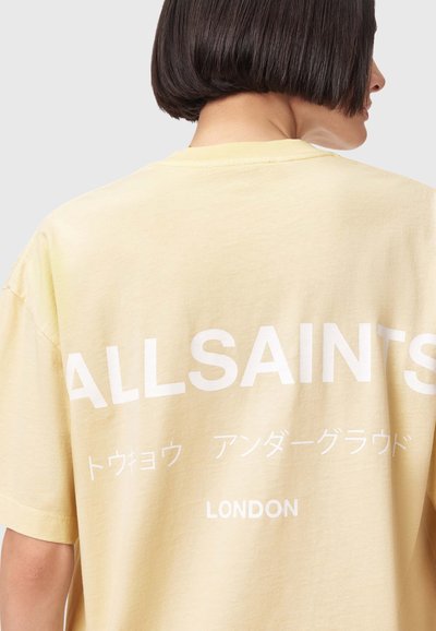 Personne aux cheveux courts et foncés portant un T-shirt jaune pâle avec « ALLSAINTS », des caractères japonais et « LONDON » imprimés en blanc dans le dos.
