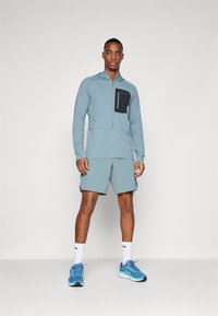 Ανοιχτό μπλε αθλητικό μπουφάν με φερμουάρ και μαύρη τσέπη στο στήθος, σε συνδυασμό με αντίστοιχα σορτς και μπλέ αθλητικά παπούτσια. Διαθέτει αναπνεύσιμο υλικό.