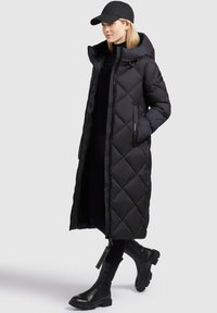 Black long puffer coat with diamond quilted pattern, zip closure, and hood. Paired with black ankle boots and a black cap.