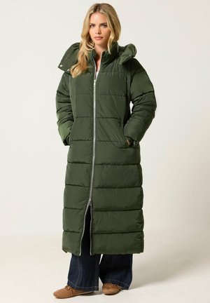 Femme portant un long manteau matelassé vert olive avec une capuche, les mains dans les poches, un jean large foncé et des chaussures plates marron.