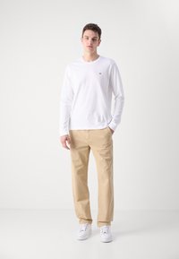 Camisa blanca de manga larga con un pequeño logo, combinada con chinos beiges y zapatillas blancas, colocada sobre un fondo neutro.