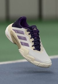 Hvide og lilla Adidas sportsko i luften over en blå tennisbane med grøn baggrund.