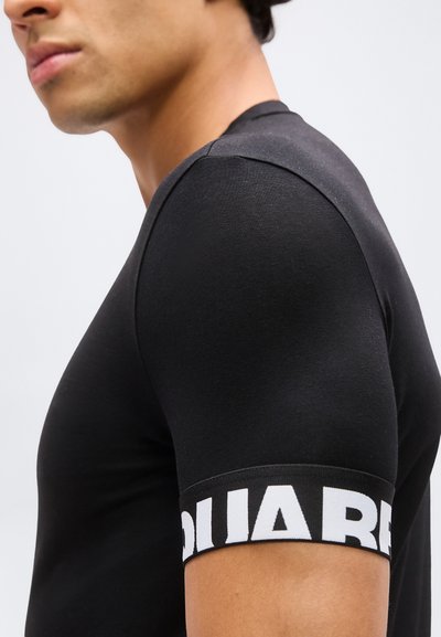 Profil d'une personne portant un t-shirt noir moulant à manches courtes avec des lettres blanches en gras sur le bracelet de la manche.