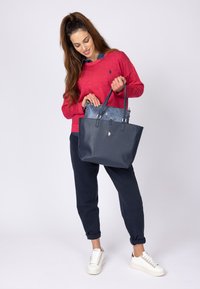 Sac en toile bleu marine avec une finition texturée, deux poignées, et une doublure intérieure à motifs. Le modèle porte un pull rouge et un pantalon bleu marine, debout dans des baskets blanches.