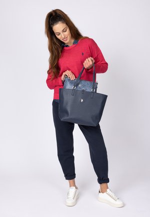 Marine tote bag med en tekstureret finish, dobbelte hanke og et mønstret indre lag. Modellen bærer en rød sweater og marineblå bukser, stående i hvide sneakers.