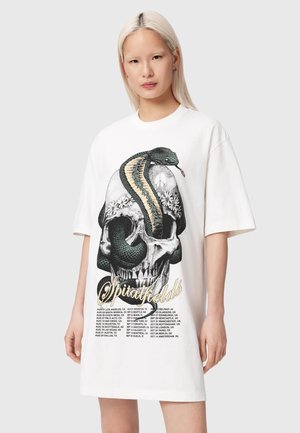 Kobieta nosząca oversize'ową białą sukienkę T-shirt z dużą grafiką czaszki z wężem i kwiatowymi detalami oraz tekstem z datami trasy koncertowej poniżej.