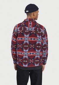 Chemise en flanelle à motifs rouges et bleus avec col évasé et manches longues, présentant des designs géométriques et une coupe décontractée, portée sur une couche noire.