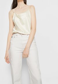 Camiseta de satén en color crema claro con escote en cowl y finas tirantes tipo spaghetti, combinada con pantalones desgastados de color beige claro.