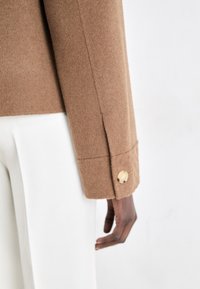 Manche de blazer en laine marron avec un détail de bouton doré au poignet, associée à un pantalon blanc. Texture douce et coupe ajustée évidente.