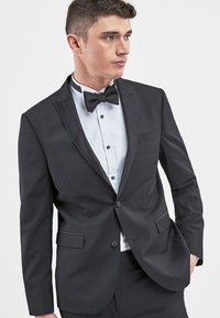 Schwarze Smokingjacke mit satinierten Revers, Ein-Knopf-Verschluss und vorderen Taschen. Getragen über einem weißen Hemd mit schwarzen Knöpfen und einer Fliege.