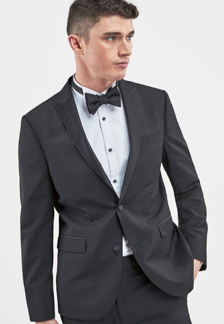 Schwarze Smokingjacke mit satinierten Revers, Ein-Knopf-Verschluss und vorderen Taschen. Getragen über einem weißen Hemd mit schwarzen Knöpfen und einer Fliege.